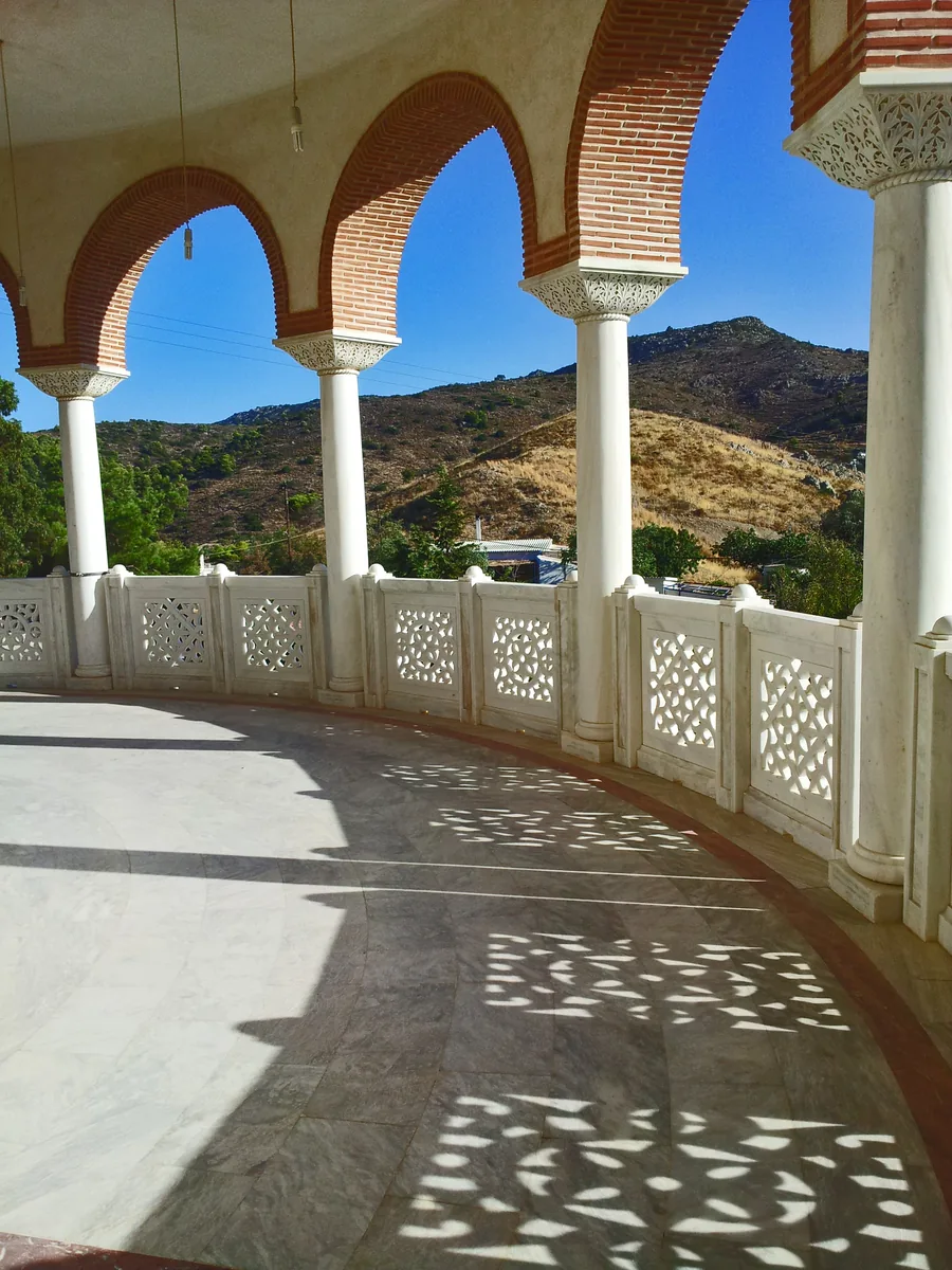 A curved open arcade of brick-trimmed arches on white marble columns, with a pierced stone balustrade and dry hills beyond.