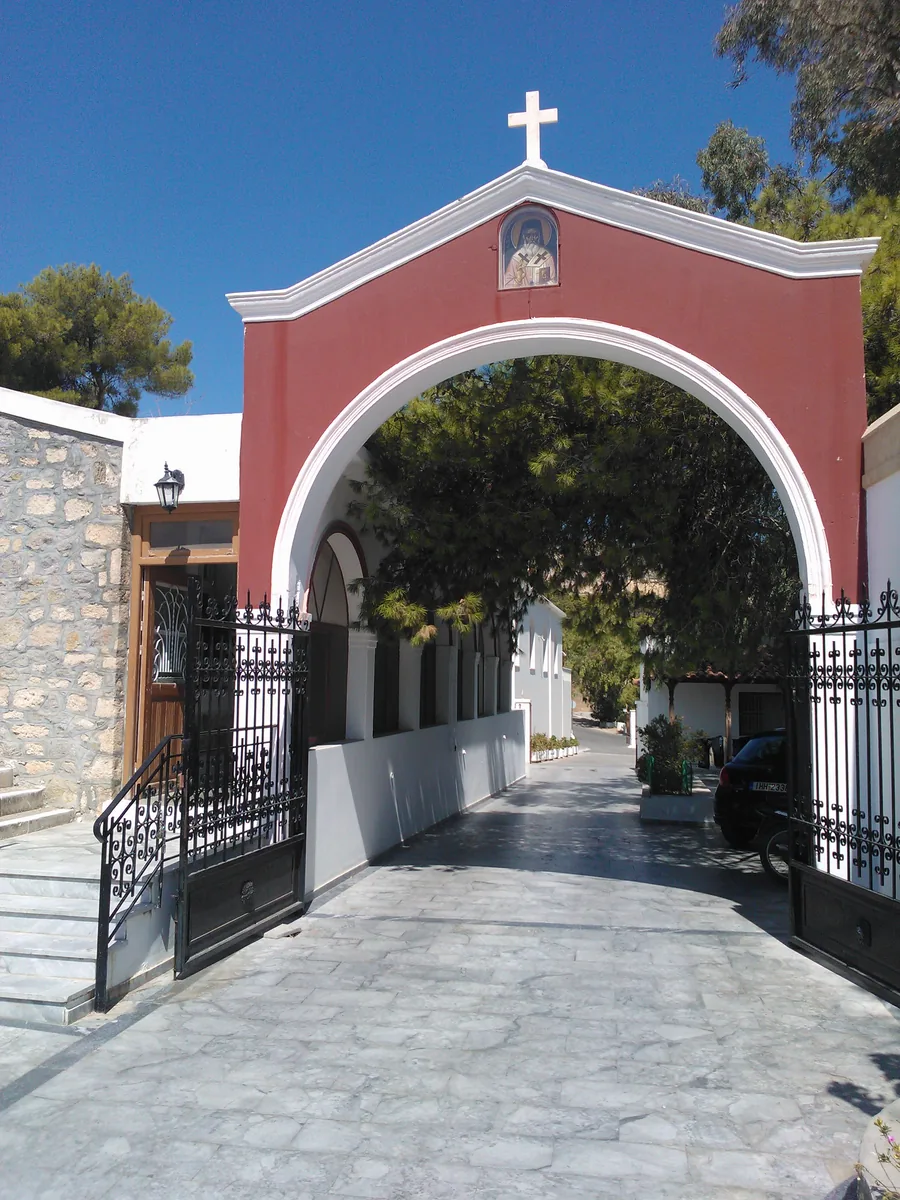 A red-painted arched gateway topped by a cross, with an icon set over the arch and iron gates standing open onto a paved drive.