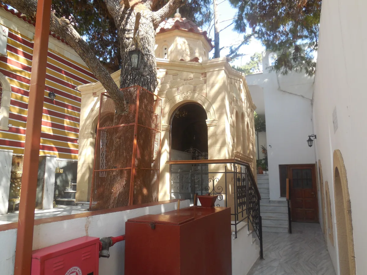 A small chapel with a tiled roof under a large pine tree, with a striped awning beside it and steps and a railing in front.