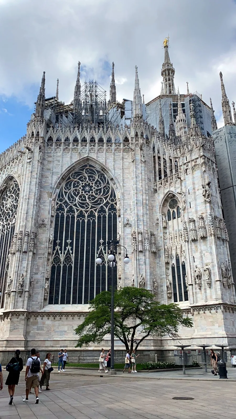 Marble Gothic pinnacles and the tracery of a large window on the flank of the cathedral, with a tree and visitors at street level.