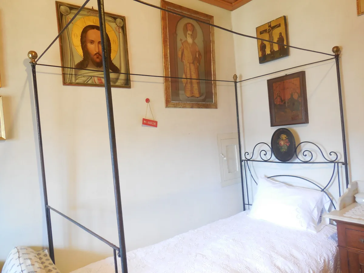 An iron four-poster bed with white linen in a whitewashed room, with icons of Christ and of a bishop hanging on the wall above it.