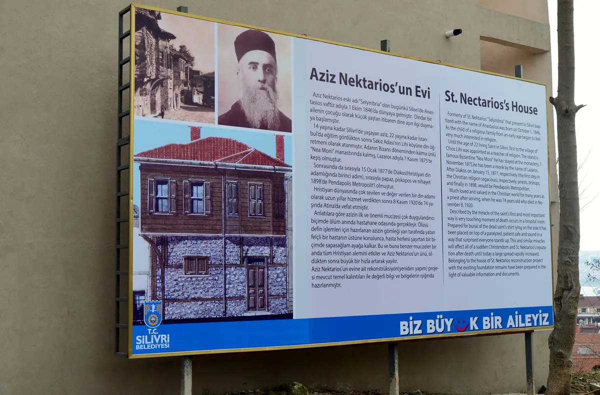 A municipal information board in Turkish and English titled St. Nectarios's House, showing an old street view, his portrait, and a drawing of the house, mounted on a wall beside an empty plot.